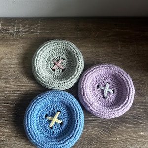 May include: Three circular crochet buttons in light gray, lavender, and blue. Each button features a recessed center with a contrasting X-shaped stitch in pink, white, or yellow thread, mimicking the appearance of a traditional four-hole button.