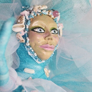 May include: A whimsical doll with a blue and white tulle dress, adorned with seashells and beads. The doll has a painted face with large eyes and a smile.