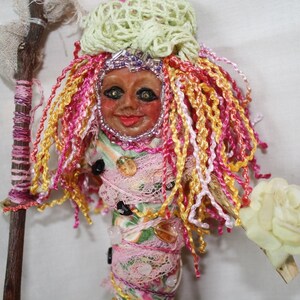 May include: A handmade doll with a colorful yarn wig, a fabric body wrapped in lace and beads, and a painted face. The doll is holding a stick with a fabric wrapped around it.