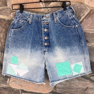 May include: High-waisted denim shorts with a button fly and a faded ombre effect. The shorts have been patched with turquoise fabric in a geometric pattern.