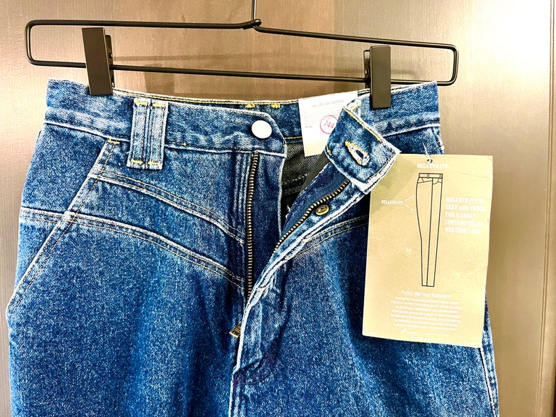 以下が含まれることがあります： ボタンとジッパー開閉の青いデニムジーンズのクローズアップ。ジーンズはリラックスしたフィット感とウエスタンスタイルのデザインです。ジーンズの図が付いたタグが添付されています。