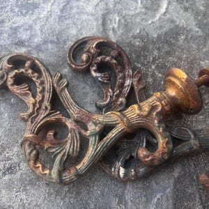 May include: A pair of ornate, rusted metal wall hooks featuring intricate scrollwork and leaf-like patterns. Each hook has a curved design with a threaded mounting screw extending from the base, showing a weathered, aged brown finish.