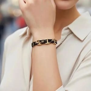 Può includere: Un bracciale caratterizzato da una serie di maglie in metallo color oro collegate da una fascia in tessuto nero con un sottile motivo color oro. Il design è composto da elementi metallici alternati a segmenti in tessuto scuro sul polso.