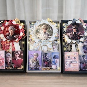 Op de afbeelding: Drie rechthoekige geschenkdozen met merchandise van anime-personages. Elke doos bevat een grote ronde badge, een decoratieve lintrozet, een sleutelhanger en diverse rechthoekige kaarten of acrylhouders met afbeeldingen van mannelijke personages.