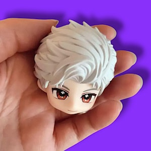 May include: A small, detailed figurine head with white hair and red eyes. The figure has a smiling expression and is held in a hand against a purple background. The figurine appears to be a character from an anime or manga.