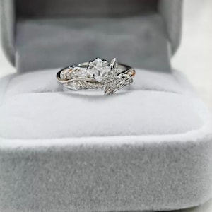 Può includere: Un anello di fidanzamento in argento con una pietra centrale a taglio marquise e delicati dettagli di foglie e viti. L'anello è presentato in un portagioie di velluto grigio, che mette in risalto il suo design intricato e la sua brillantezza.