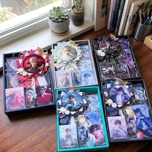 May include: A collection of gift boxes, each containing photographs, badges, and keyrings. The boxes are decorated with large circular pins featuring a person's image and ribbons. The boxes are in various colours, including black and teal, and are arranged on a wooden surface.