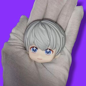 Pode incluir: Uma pequena cabeça de figura em estilo anime com cabelo cinza claro e olhos azuis. A figura tem uma expressão neutra com uma boca pequena e vermelha. A cabeça é segurada em uma mão enluvada contra um fundo roxo.