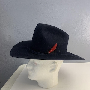 May include: A dark navy blue cowboy hat with a wide brim and a decorative red feather. The hat is displayed on a white mannequin head. The hat has a black band around the crown.