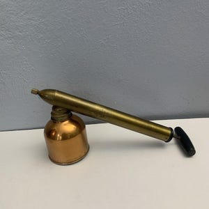 May include: A vintage brass and copper sprayer with a cylindrical copper base and a long brass nozzle. The sprayer has a black handle and a small nozzle at the end. The sprayer is likely used for gardening or other liquid applications.