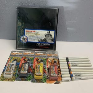 May include: A collection of art supplies including a black Koh-I-Noor Rapidograph set, several Trans Mix bottles, and a set of Koh-I-Noor pens with colored tips. The Rapidograph set features a wizard illustration.