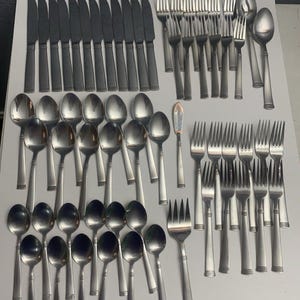 May include: A collection of stainless steel silverware, including knives, forks, spoons, and a butter knife. The silverware has a simple, modern design with a brushed finish. The arrangement suggests a complete set for multiple place settings.