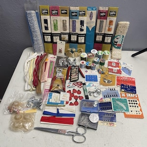 May include: A collection of sewing supplies, including Talon zippers, spools of thread, snaps, scissors, and various notions. The items are arranged on a white surface, with a variety of colors and textures.