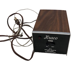May include: A vintage Kraco 100 DC solid state power converter with a brown wood-grain top and black front panel. The front panel has the Kraco logo, a switch, and a red indicator light. A brown power cord is attached.