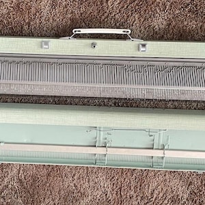 May include: Vintage knitting machine in light green and gray. The machine has a handle and several buttons and dials. The machine is open, with the top and bottom sections separated, revealing the internal components. The machine is designed for creating knitted items.