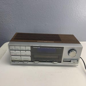 May include: A vintage Soundesign AM-FM electronic dual alarm clock radio. The radio has a silver face with a digital display and a wood-grain top. The controls include buttons for time, alarm, and radio functions.