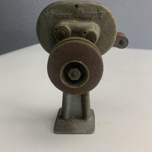 May include: A vintage gray bench grinder with a brown grinding wheel. The grinder has a red handle and is mounted on a rectangular base. The name "PARKER" is visible on the top of the grinder.