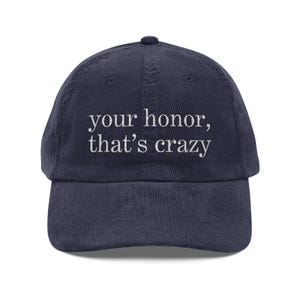 May include: Navy blue corduroy baseball cap with the embroidered text "your honor, that's crazy" in white. The cap has a curved brim and a small button on top. A casual accessory.