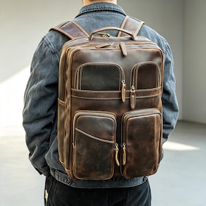 May include: A dark brown leather backpack featuring a rectangular shape, multiple exterior zippered pockets, a top handle, and adjustable shoulder straps. The bag has visible stitching details and metal zipper pulls throughout the design.