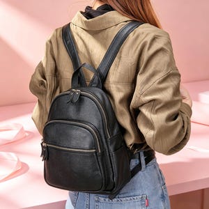 Könnte beinhalten: Ein schwarzer Lederrucksack mit runder Form, mit einer Reißverschlusstasche vorne und zwei Reißverschlüssen oben. Der Rucksack hat verstellbare schwarze Träger und einen oberen Griff. Die Tasche wird über den Schultern getragen.
