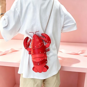 May include: A bright red lobster-shaped crossbody bag featuring a segmented shell design with silver-toned metal studs, two large claws, and a thin shoulder strap. The bag is worn over a plain white t-shirt and light-coloured trousers, showcasing its structure.