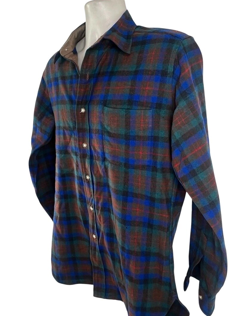 May include: A long-sleeve button-down shirt featuring a plaid pattern in shades of blue, green, brown, and black. The shirt has a pointed collar, a single chest pocket on the left side, and light-colored buttons running down the front placket.