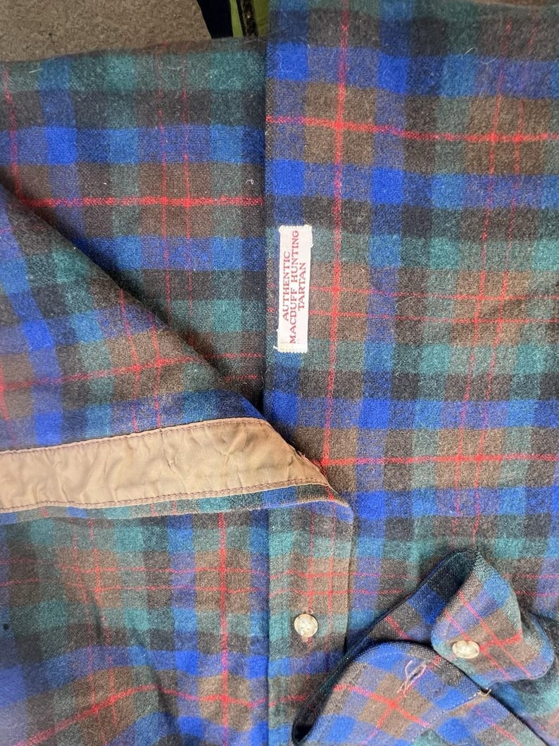 May include: A long-sleeve shirt featuring a Macduff Hunting tartan pattern in shades of blue, green, black, and red. The shirt includes a tan inner collar lining, light-colored buttons, and a white fabric label reading AUTHENTIC MACDUFF HUNTING TARTAN.