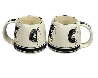 Puede incluir: Dos tazas de cerámica blanca con diseños negros de figuras estilizadas. Las tazas tienen bandas negras alrededor de la parte superior e inferior.