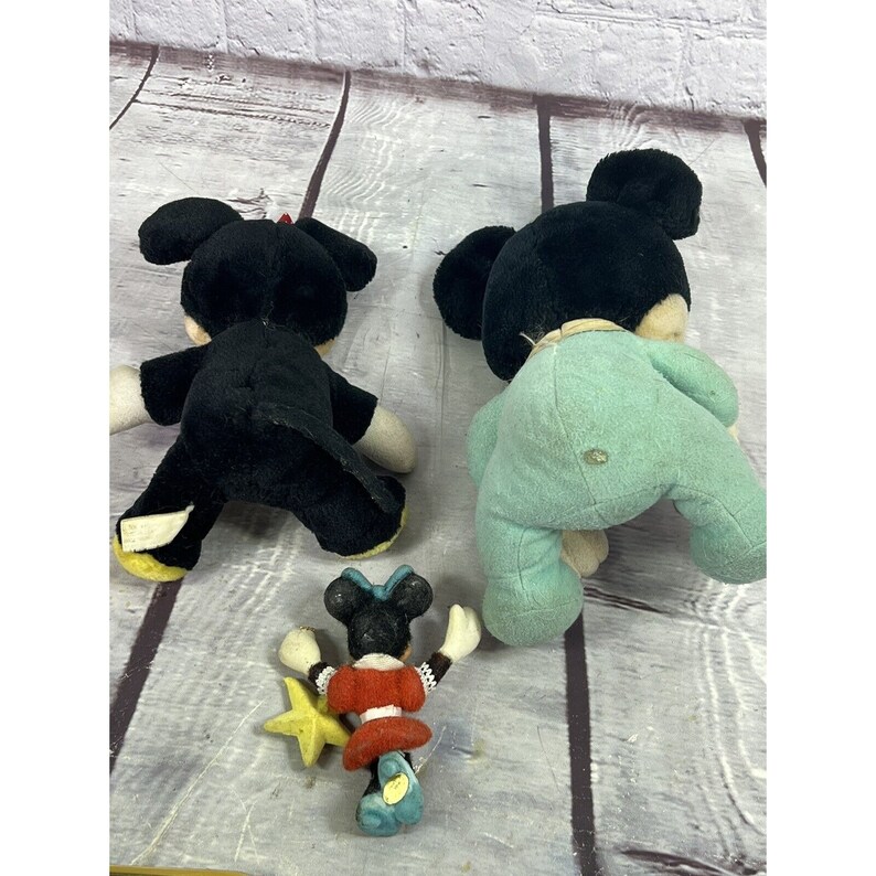 May include: Three plush Mickey Mouse toys. A black Mickey Mouse with a red bow tie, a black Mickey Mouse wearing a light blue onesie, and a smaller red and black Minnie Mouse holding a yellow star.