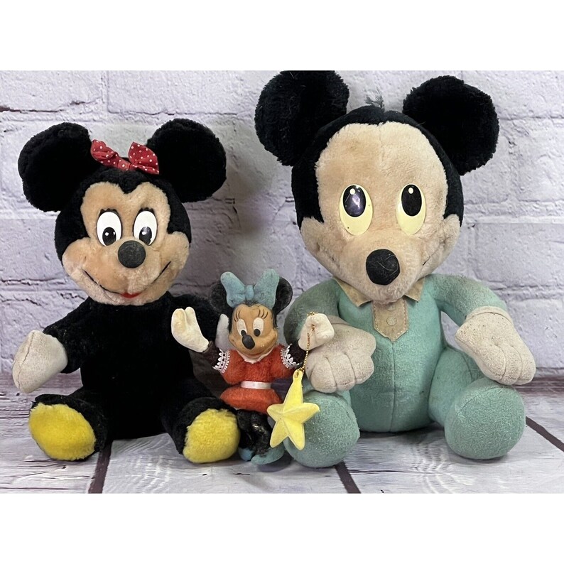 May include: Three plush Mickey Mouse toys. A large black and white Mickey Mouse with a red bow, a smaller black and white Mickey Mouse with a red dress and a yellow star, and a large green and white Mickey Mouse wearing a blue shirt.