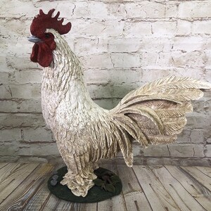 May include: A detailed rooster statue with a cream-colored body, a red comb and wattle, and a beige tail. The rooster stands on a green base with small rocks and leaves. The statue is set against a white brick wall and wooden floor.