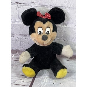 May include: A black plush Minnie Mouse toy with a red polka dot bow. The toy has a brown face with large eyes and a wide smile. The toy is sitting with its legs crossed and its arms outstretched.
