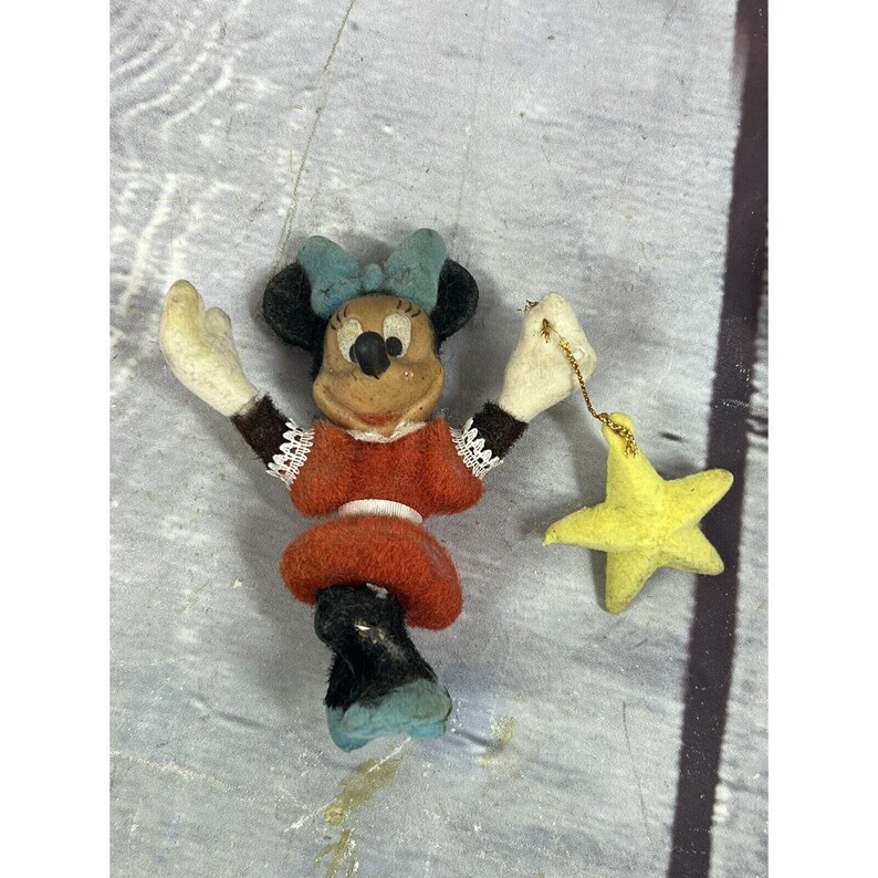 May include: A vintage plush Minnie Mouse figurine with a red dress, black shoes, and a blue bow. She is holding a yellow star with a gold string.