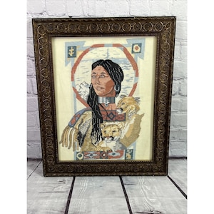 Puede incluir: Obra de punto de cruz enmarcada que representa a una mujer nativa americana con el pelo largo y negro, que lleva un collar de cuentas y ropa tradicional. Está sosteniendo dos pumas, uno a cada lado de ella. La obra está ambientada en un fondo de montañas y cielo azul.