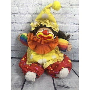 May include: A Cabbage Patch Kid doll dressed as a clown. The doll has brown hair, a red nose, and a yellow and red clown costume with a rainbow trim. The doll is sitting on a wooden floor.