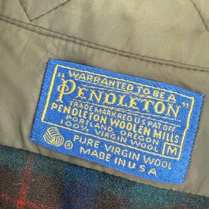 May include: A blue rectangular clothing label with yellow text stitched onto a tan quilted lining. The label reads: Warranted to be a Pendleton, Pendleton Woolen Mills, Portland, Oregon, 100% Virgin Wool, Pure Virgin Wool, Made in U.S.A.