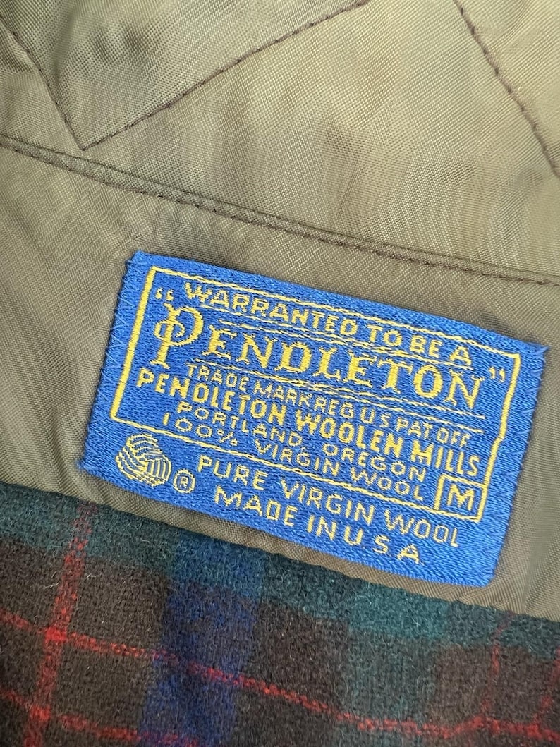 May include: A blue rectangular clothing label with yellow text stitched onto a tan quilted lining. The label reads: Warranted to be a Pendleton, Pendleton Woolen Mills, Portland, Oregon, 100% Virgin Wool, Pure Virgin Wool, Made in U.S.A.