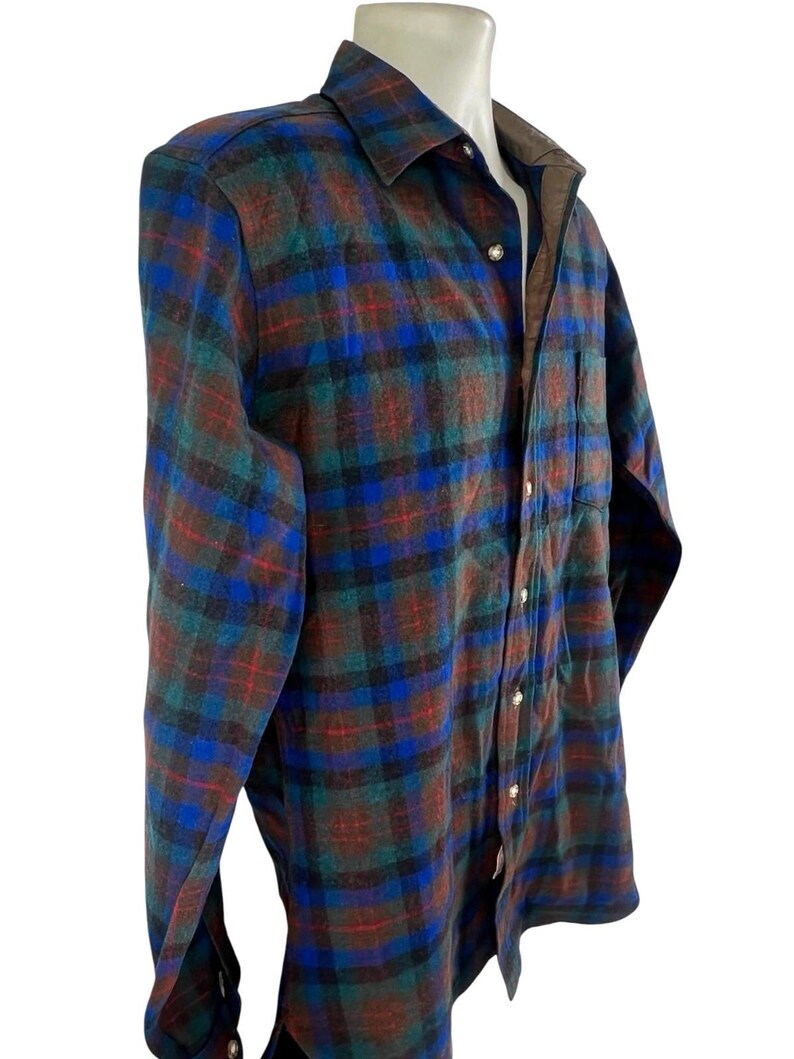 May include: A long-sleeve button-down shirt featuring a plaid pattern in shades of blue, green, and brown. The shirt has a pointed collar, a single chest pocket, and light-colored buttons running down the front placket. The interior collar lining is brown.