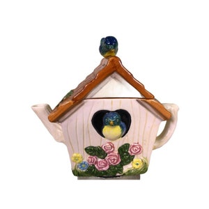 May include: A white ceramic teapot shaped like a birdhouse with a brown roof. The teapot has a blue bird on the roof and a blue bird in the heart-shaped opening. The teapot is decorated with pink roses and green leaves.