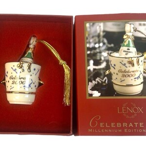 May include: A white porcelain ornament depicting a bottle of champagne in an ice bucket. The ornament is decorated with gold trim and blue and white confetti. The text "Celebrate 2000" is printed on the ornament. The ornament is packaged in a red box with gold trim and the text "Lenox Celebrate 2000 Millennium Edition Ornament".