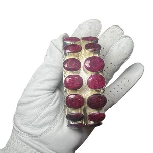 May include: A silver bracelet featuring large, oval-cut, deep red gemstones. The bracelet is held in a white glove, showcasing the polished metal and the vibrant color of the stones. The bracelet is a statement piece.