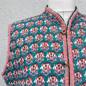 Op de afbeelding: Een mouwloos gewatteerd vest met een groenblauwe achtergrond en een herhalend bloemenpatroon in rood, wit en blauw. Het kledingstuk heeft een hoge kraag, roze fluwelen biezen langs de randen en drie ronde knopen met bloemmotieven aan de voorzijde.