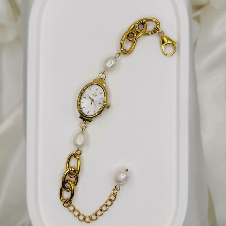 May include: Gold-toned bracelet watch featuring an oval watch face with a white dial. The bracelet combines large chain links with freshwater pearls. A lobster clasp secures the watch. The watch is displayed on a white surface.