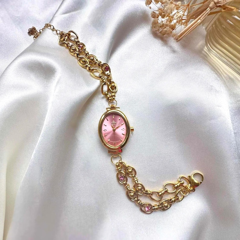 May include: Gold-toned watch with a pink oval face and a delicate chain bracelet. The watch features a small crown emblem on the face and is adorned with pink gemstones. The chain has a double-strand design.