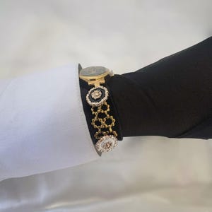 May include: A gold-toned watch with a black face and a decorative bracelet. The bracelet features black and white circular accents with gold detailing. The watch is worn over a black sleeve and a white shirt cuff.