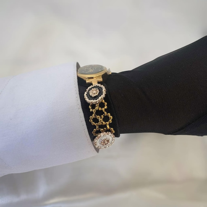 May include: A gold-toned watch with a black face and a decorative bracelet. The bracelet features black and white circular accents with gold detailing. The watch is worn over a black sleeve and a white shirt cuff.