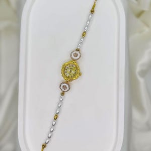 May include: A gold-toned wristwatch with a cream-colored face and delicate floral accents. The watch is attached to a bracelet with alternating pearl and gold-colored beads. The watch face displays the numbers 1 through 12.