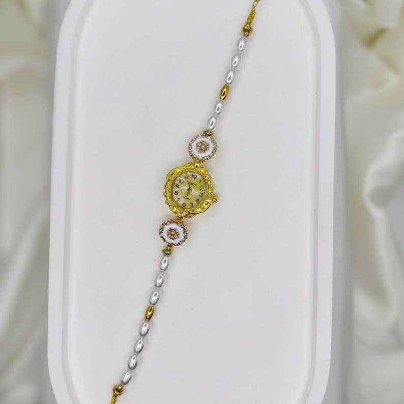 May include: A gold-toned wristwatch with a cream-colored face and delicate floral accents. The watch is attached to a bracelet with alternating pearl and gold-colored beads. The watch face displays the numbers 1 through 12.
