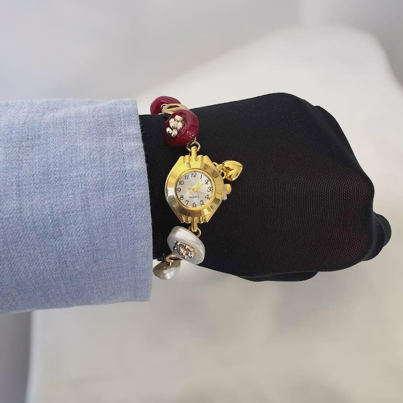 May include: Gold-toned quartz watch with a white face and gold hands. The watch is attached to a red and white beaded bracelet. The watch face has the numbers 1 through 12. The bracelet is attached to a gold-toned clasp.