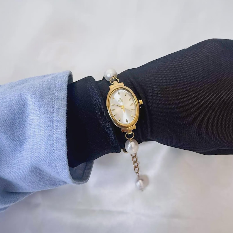 May include: Gold-toned oval wristwatch with a white face and delicate hands. The watch features a pearl-adorned chain and a pearl at each end of the watch face. The watch is on a black surface, with a light blue sleeve in the background.